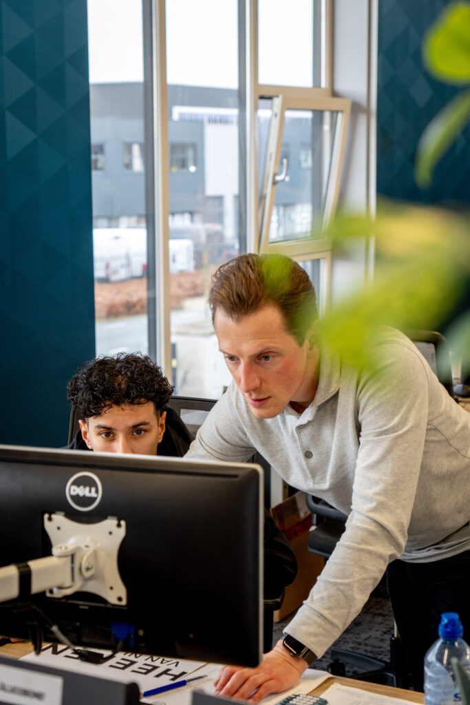 Begeleider helpt een stagiair bij computerwerk op kantoor en kijkt samen mee op het scherm.