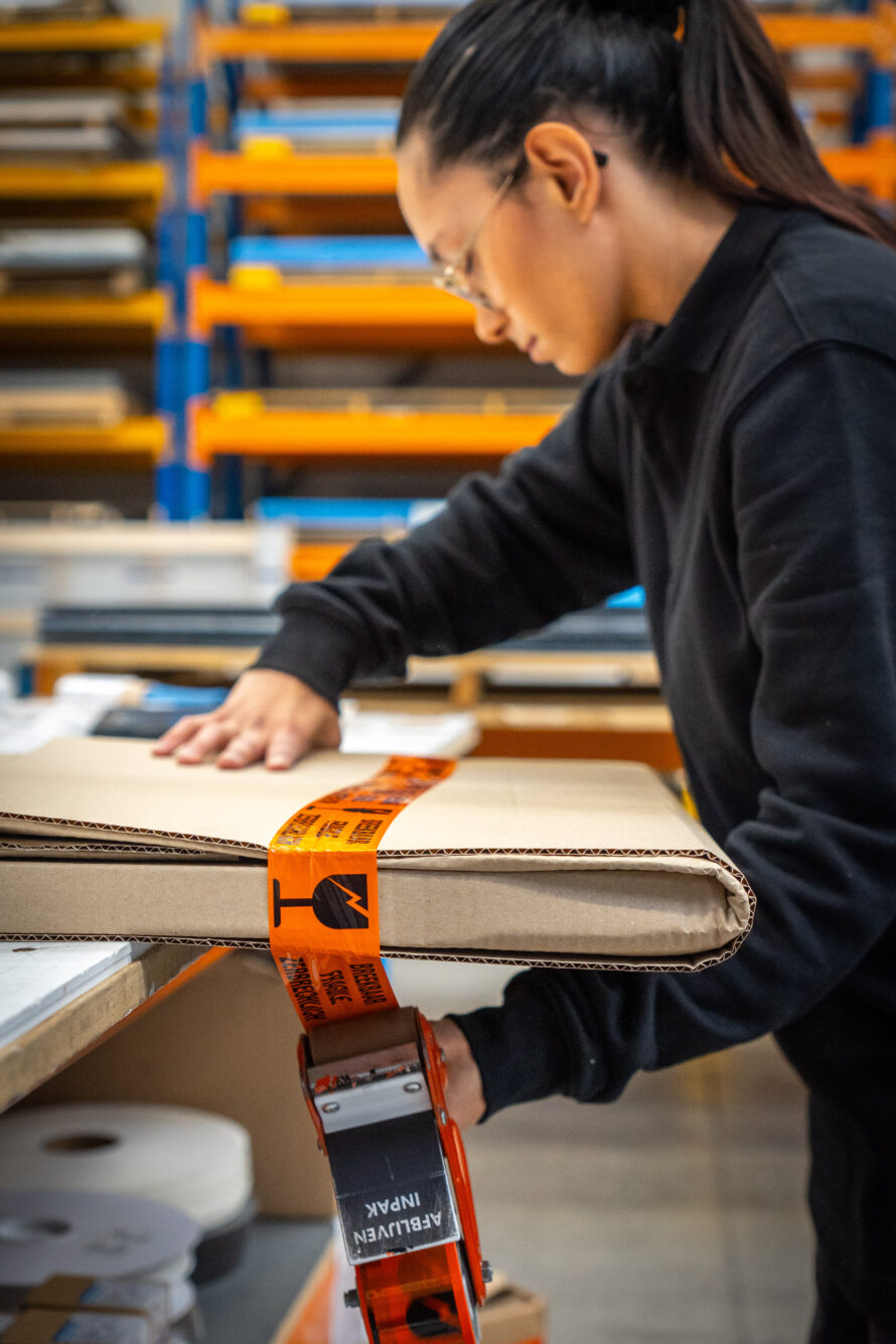 Medewerker plakt beschermende tape op een plexiglas product tijdens het inpakken op de werkvloer.