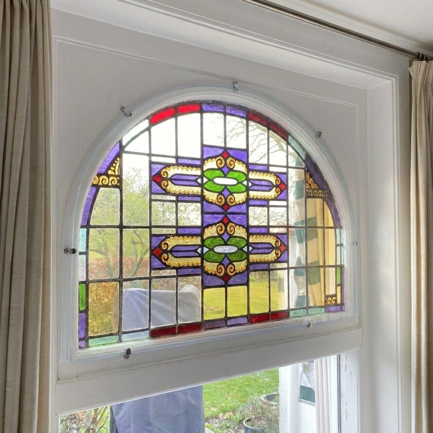 Voorzetraam voor glas-in-lood in boogvormig bovenlicht, gemonteerd aan de binnenzijde van een wit houten kozijn.