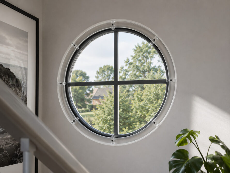 Rond raam in een lichte kamer, met uitzicht op groen gebladerte en een plant zichtbaar in de hoek van het interieur.