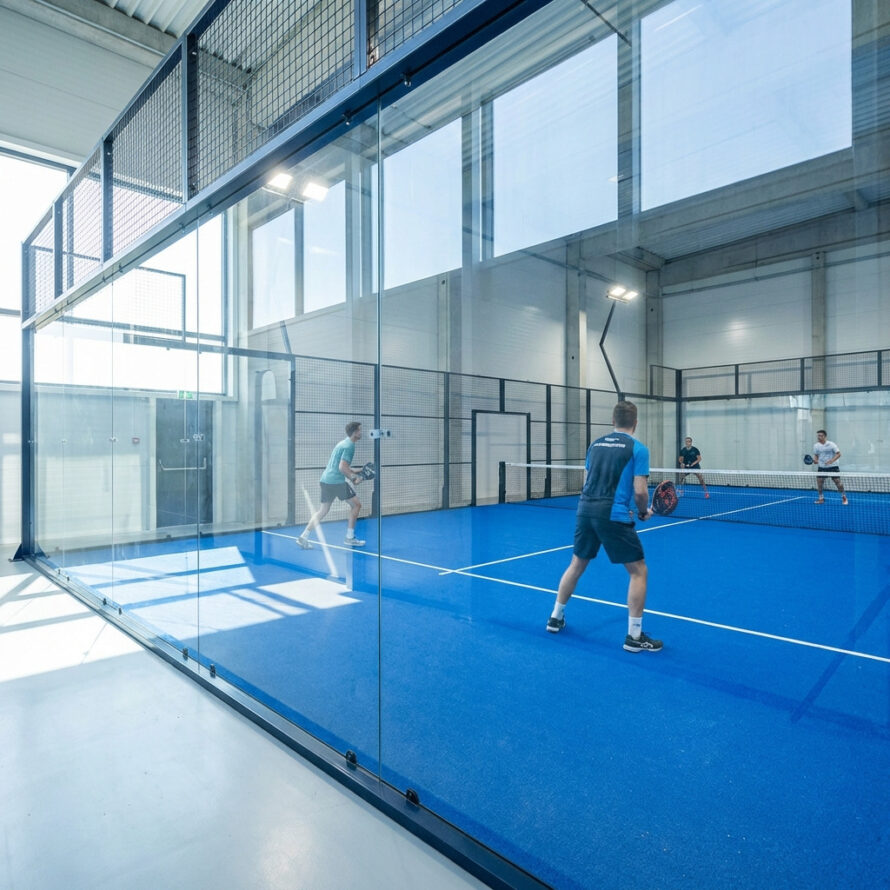 Zijaanzicht van polycarbonaat padelwand met metalen bevestigingsklemmen in indoor sporthal