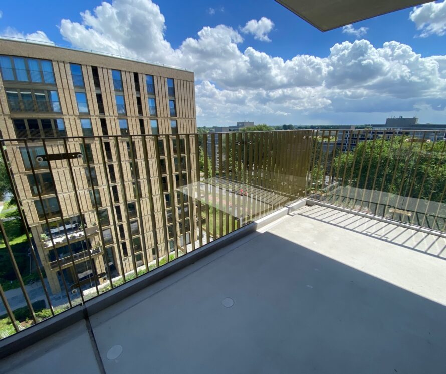 Balkon met glazen balustrade en metalen spijlen, met uitzicht op omliggende appartementen en groen onder een blauwe lucht.