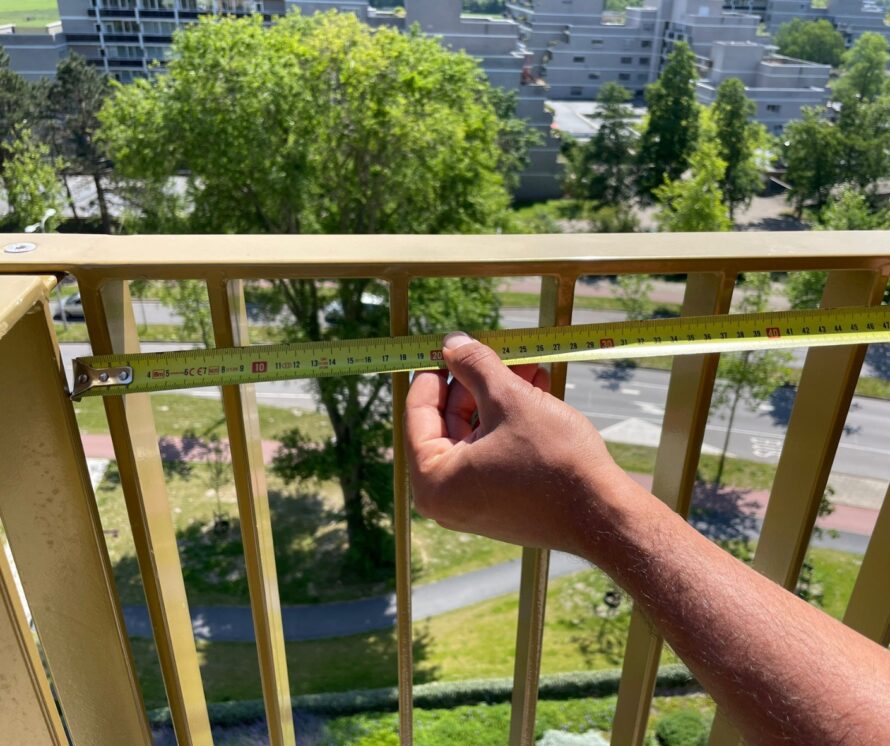 Detail van een metalen balkonhek waarbij met een rolmaat de afstand tussen de verticale spijlen wordt gemeten.