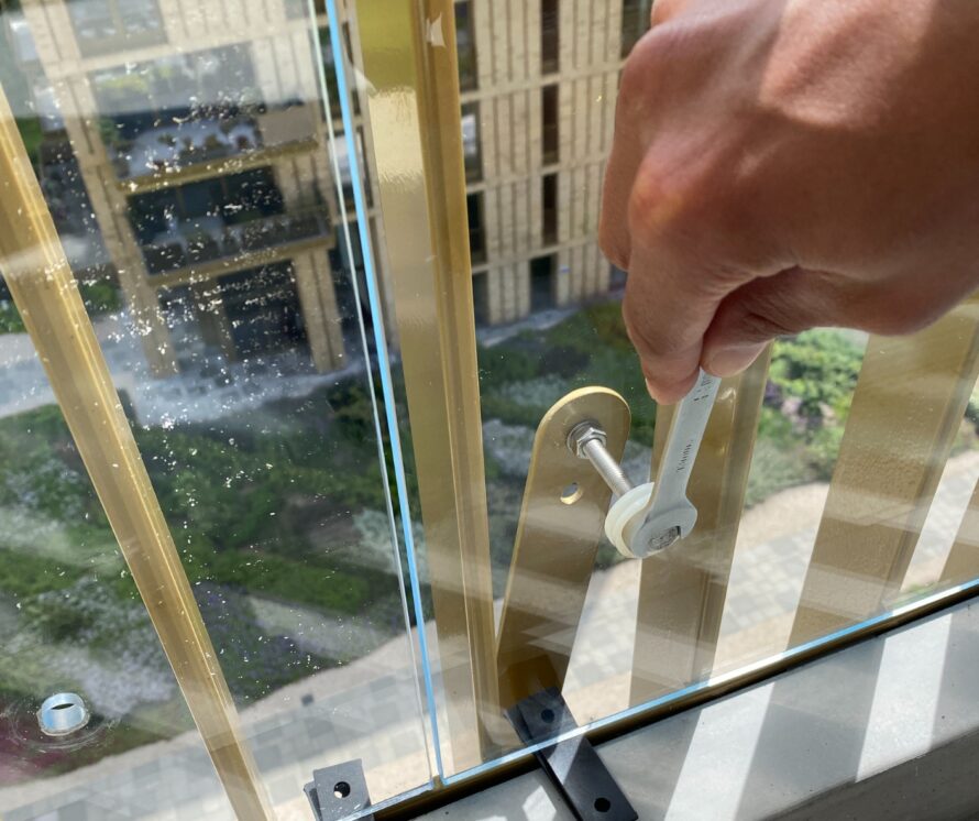 Close-up van bevestiging van een glazen balkonpaneel aan een metalen hek, waarbij een bout met een sleutel wordt aangedraaid.
