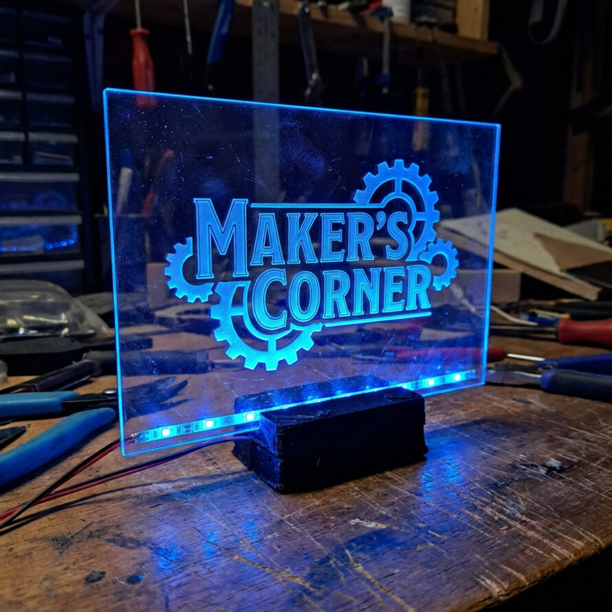 Edge-lit plexiglas LED bord met gravure “Makers Corner”, blauw verlicht en geplaatst op zwarte standaard.