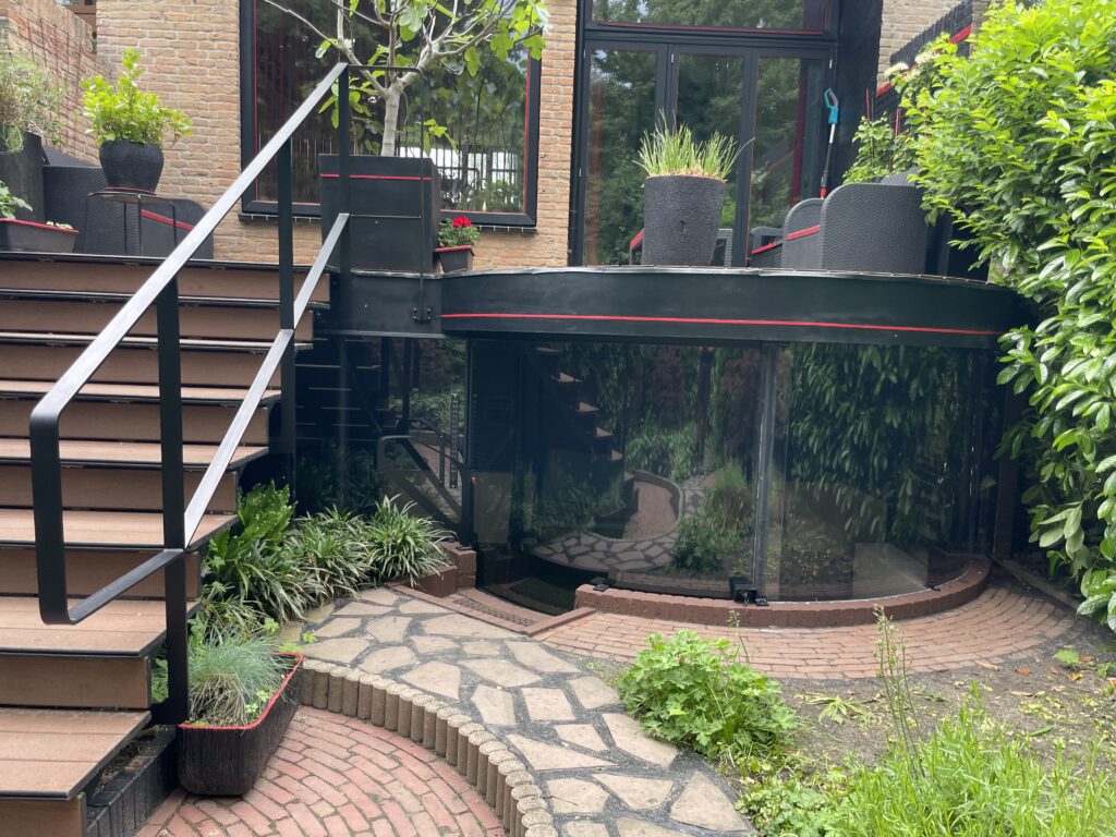 Gebogen plexiglas serre geïntegreerd in een tuin met stenen pad en beplanting.