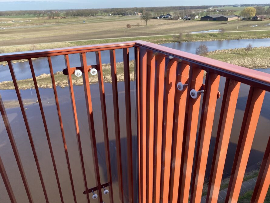 Close-up van een metalen hoekrailing van een uitkijkplatform met water op de achtergrond.