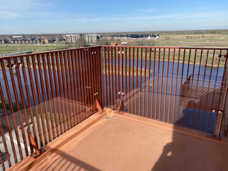 Hoek van een uitkijkplatform met metalen spijlenrailing en uitzicht over het water.