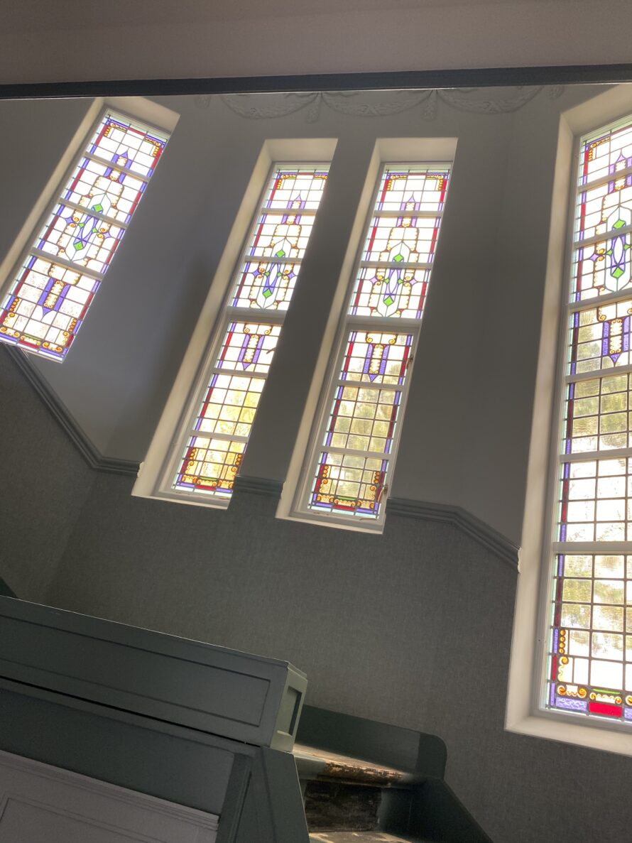 Hoge glas-in-loodramen in een lichte hal met schuine wand.