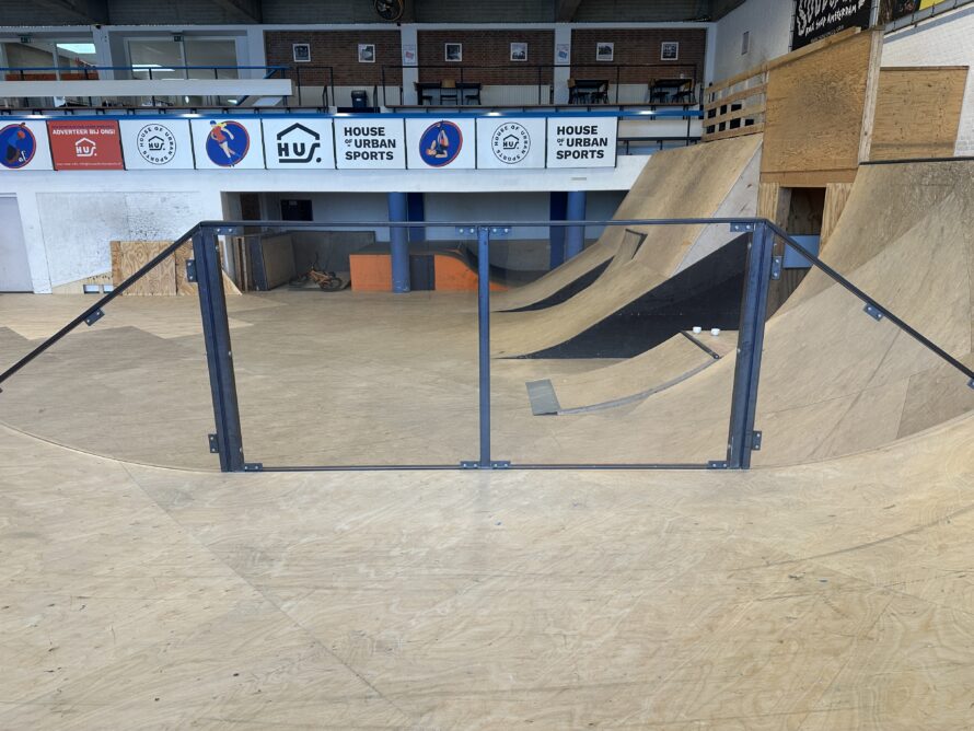 Indoor skatepark met een metalen handrail op een betonnen vloer en een houten ramp op de achtergrond.