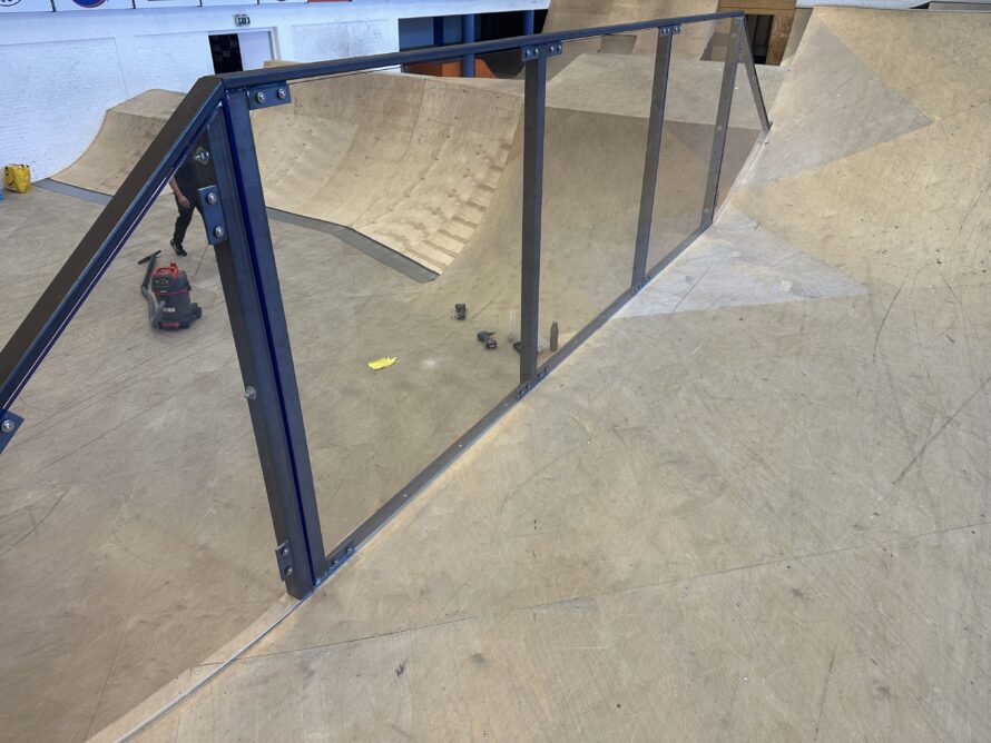 Transparante afscherming van helder polycarbonaat gemonteerd in donkerblauwe stalen frames langs een skatepark ramp.
