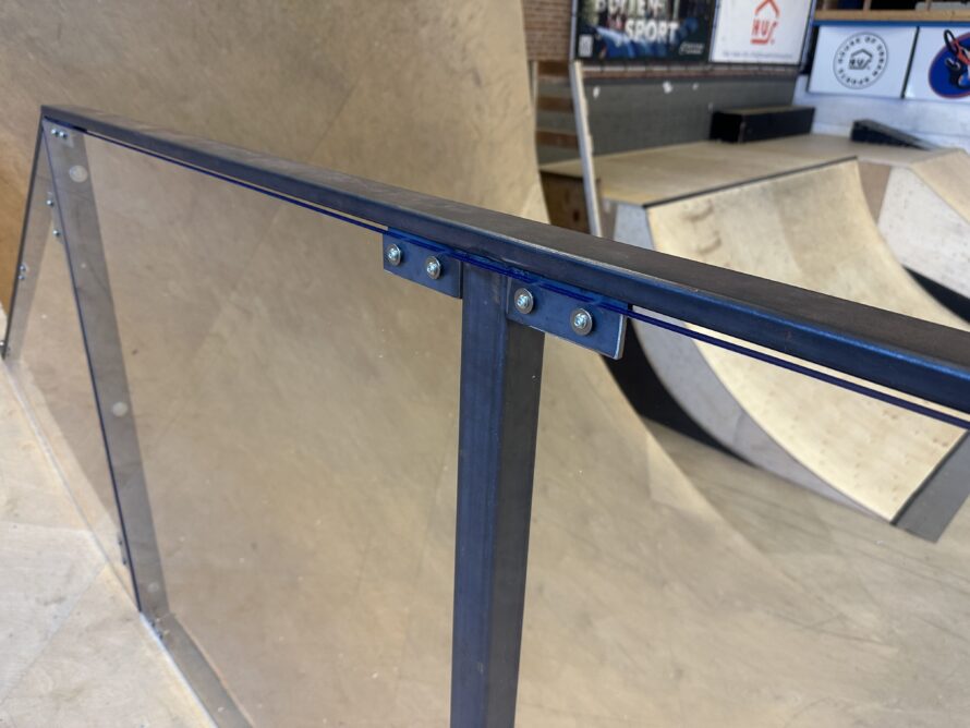 Detail van een transparante polycarbonaatplaat bevestigd in een blauw stalen frame met metalen bevestigingspunten langs een ramp.