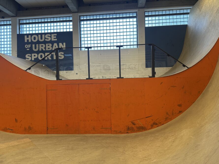 Overzicht van een skatepark met een oranje ramp en transparante polycarbonaat afscherming langs de rand.