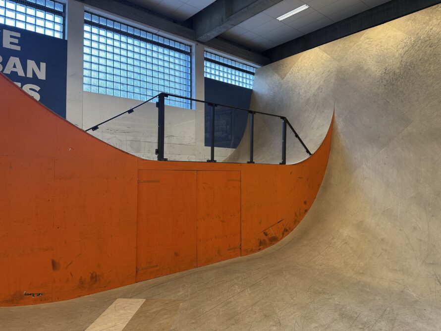 Binnenruimte van een skatepark met een gebogen betonnen ramp en een feloranje leuning langs de rand; op de achtergrond zijn ramen en metalen constructies zichtbaar.